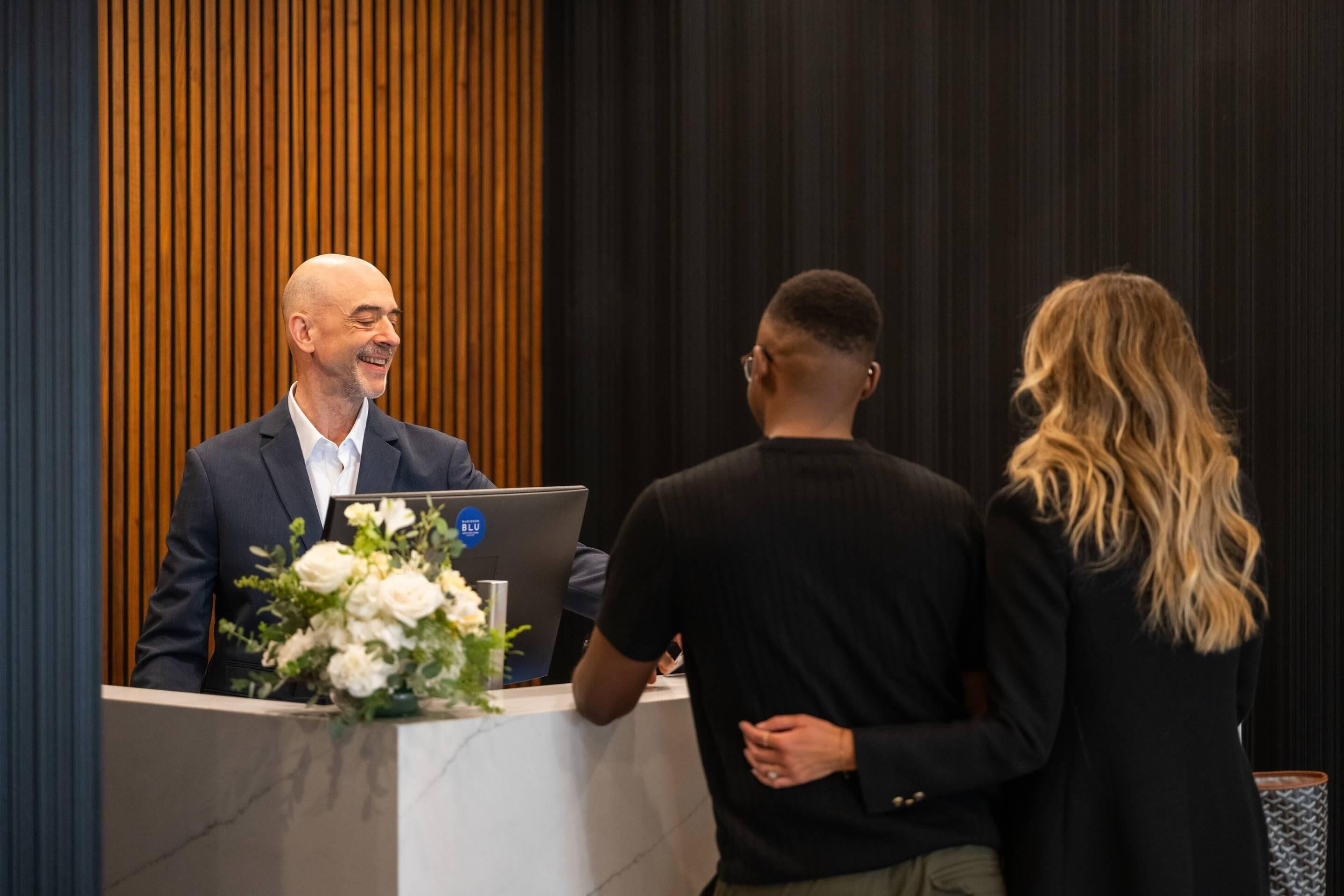A man in a suit standing at a podium with a laptop and a floral arrangement, addressing two individuals.