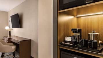 Ein Hotelzimmer mit Schreibtisch, Stuhl, flachbildfernseher an der Wand, einer Kaffeestation inklusive Kaffeemaschine, Tassen und einer Auswahl an Gebäck.