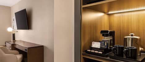 A hotel room with a desk, a chair, a flat-screen TV mounted on the wall, a coffee station with a coffee maker, cups, and a tray of pastries.