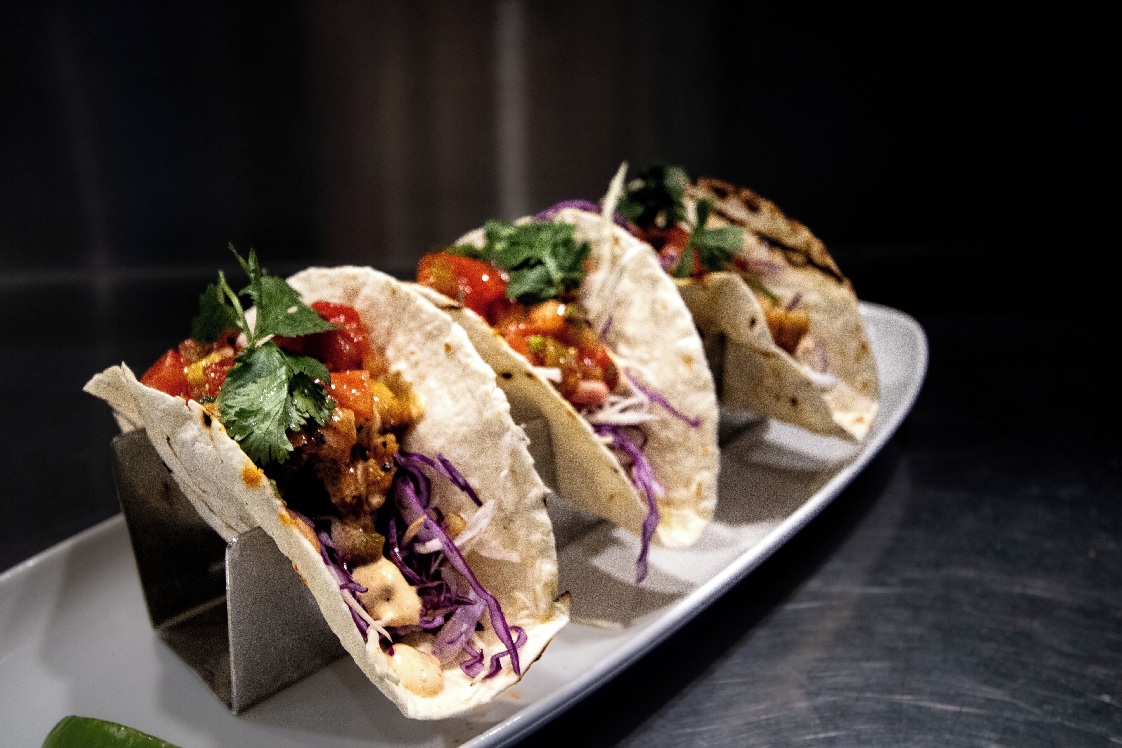Tre tacos på en hvid tallerken mod en mørk baggrund.