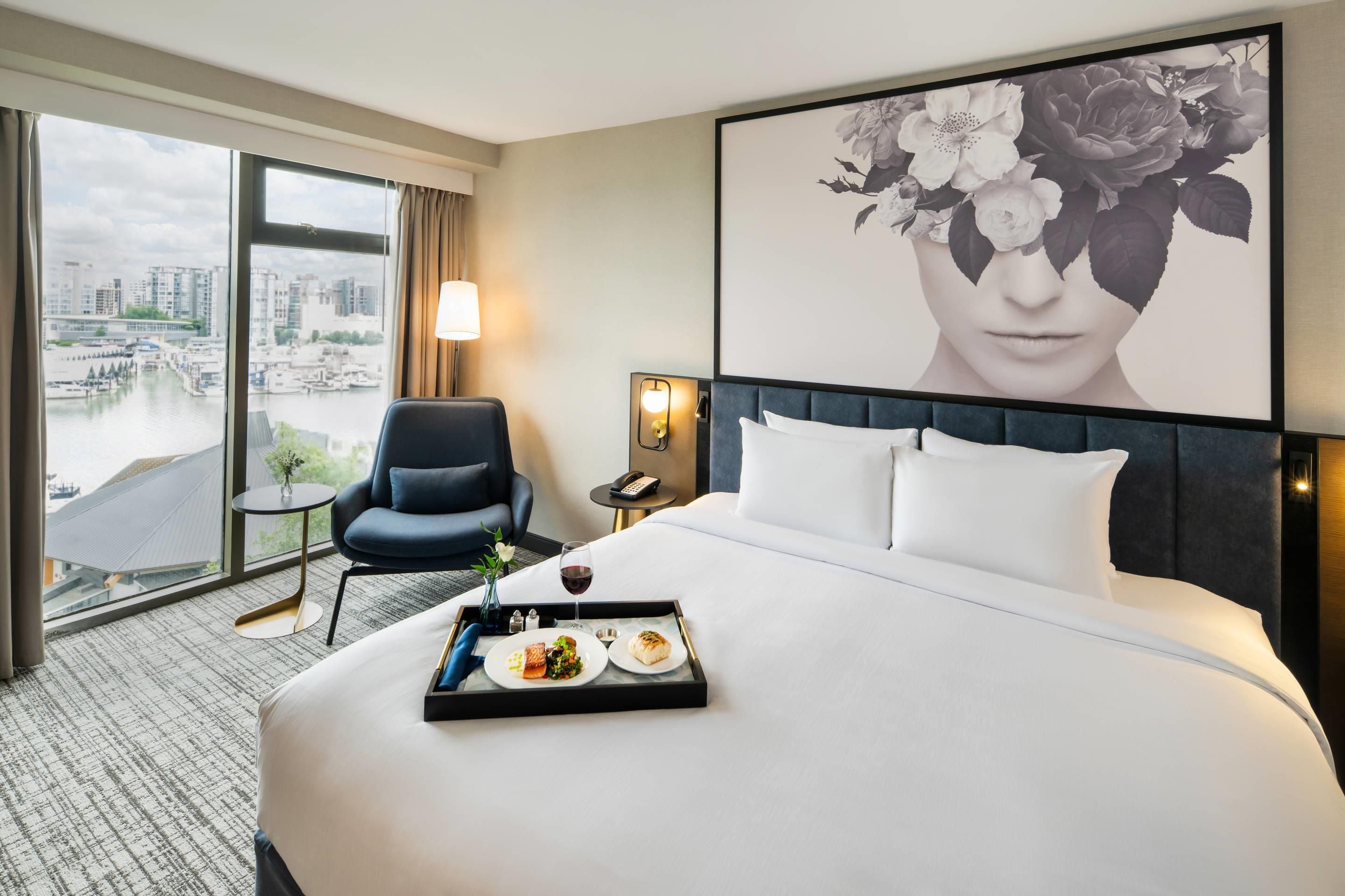 A hotel room with a large bed, a chair, a small table with a tray of food, a lamp, and a large window with a city view.