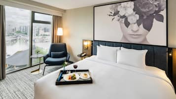 A hotel room with a large bed, a chair, a small table with a tray of food, a lamp, and a large window with a city view.