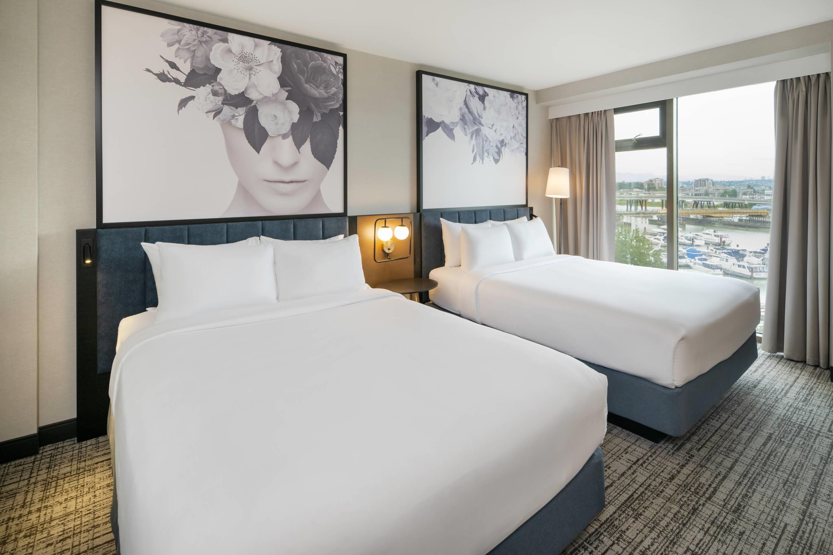 A hotel room with two beds, a large window with a view of a cityscape, and artwork on the walls.