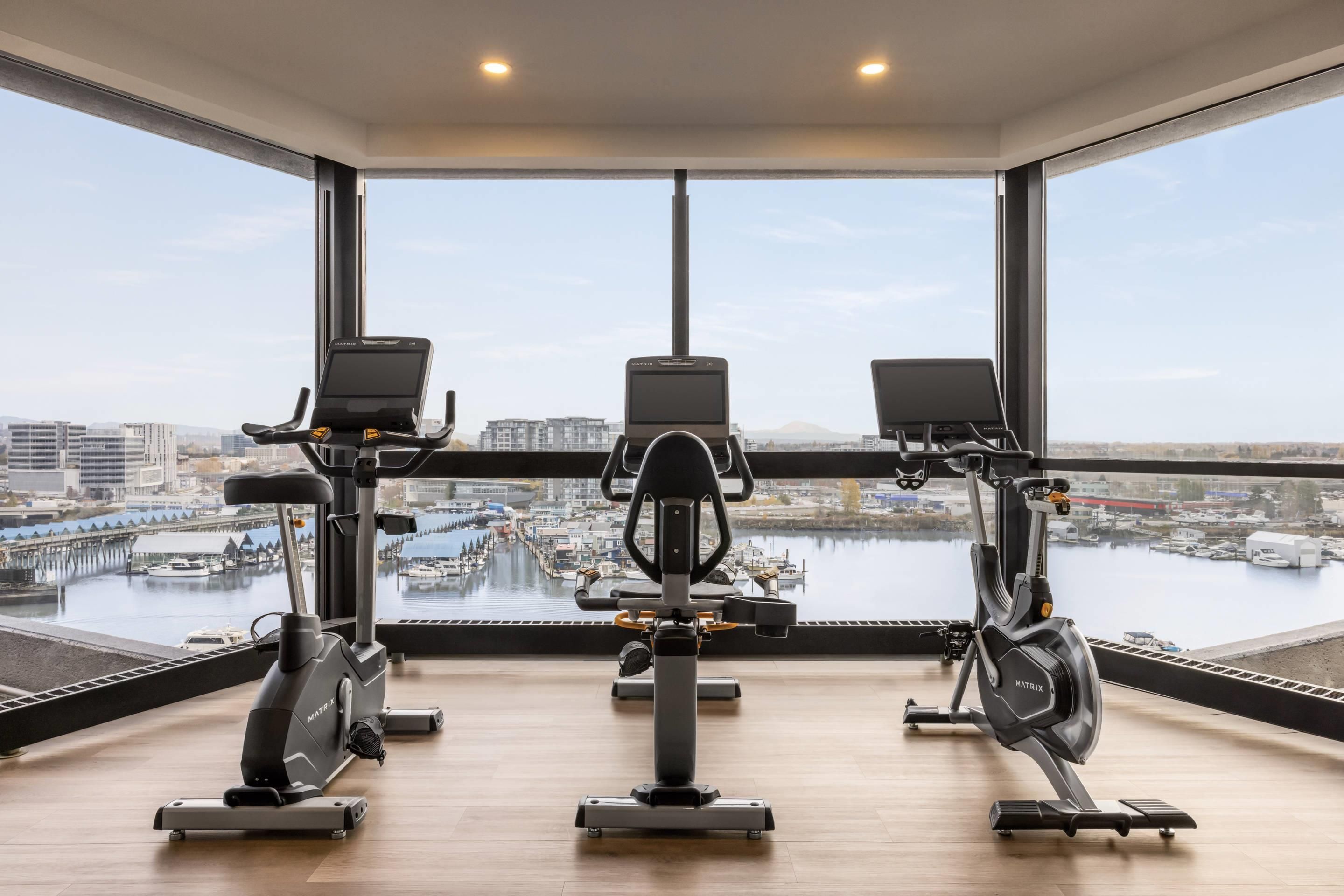 A gym with three stationary bikes, large windows, and a view of a marina.