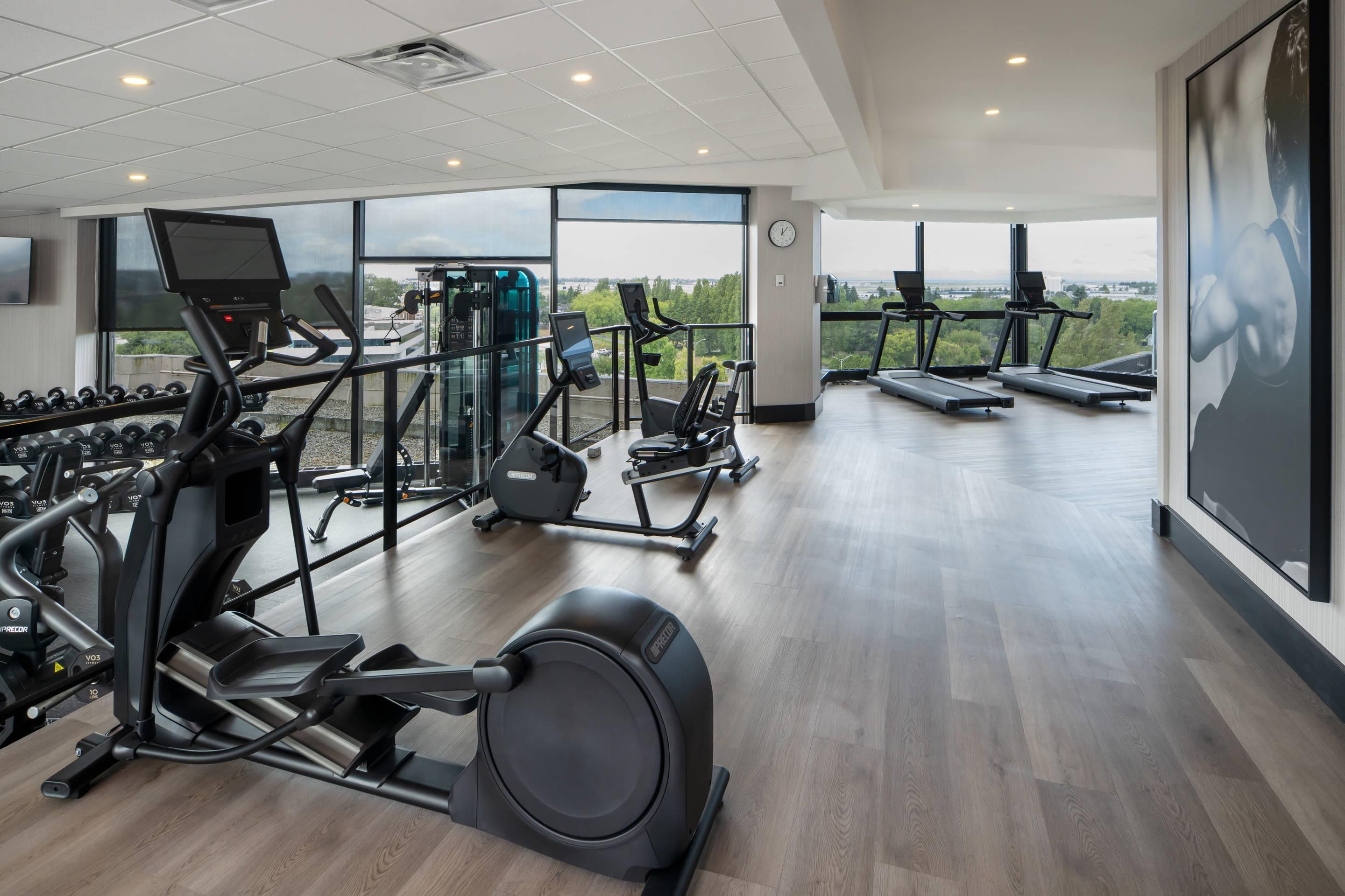 A modern gym with cardio machines, free weights, and a large window offering a city view.
