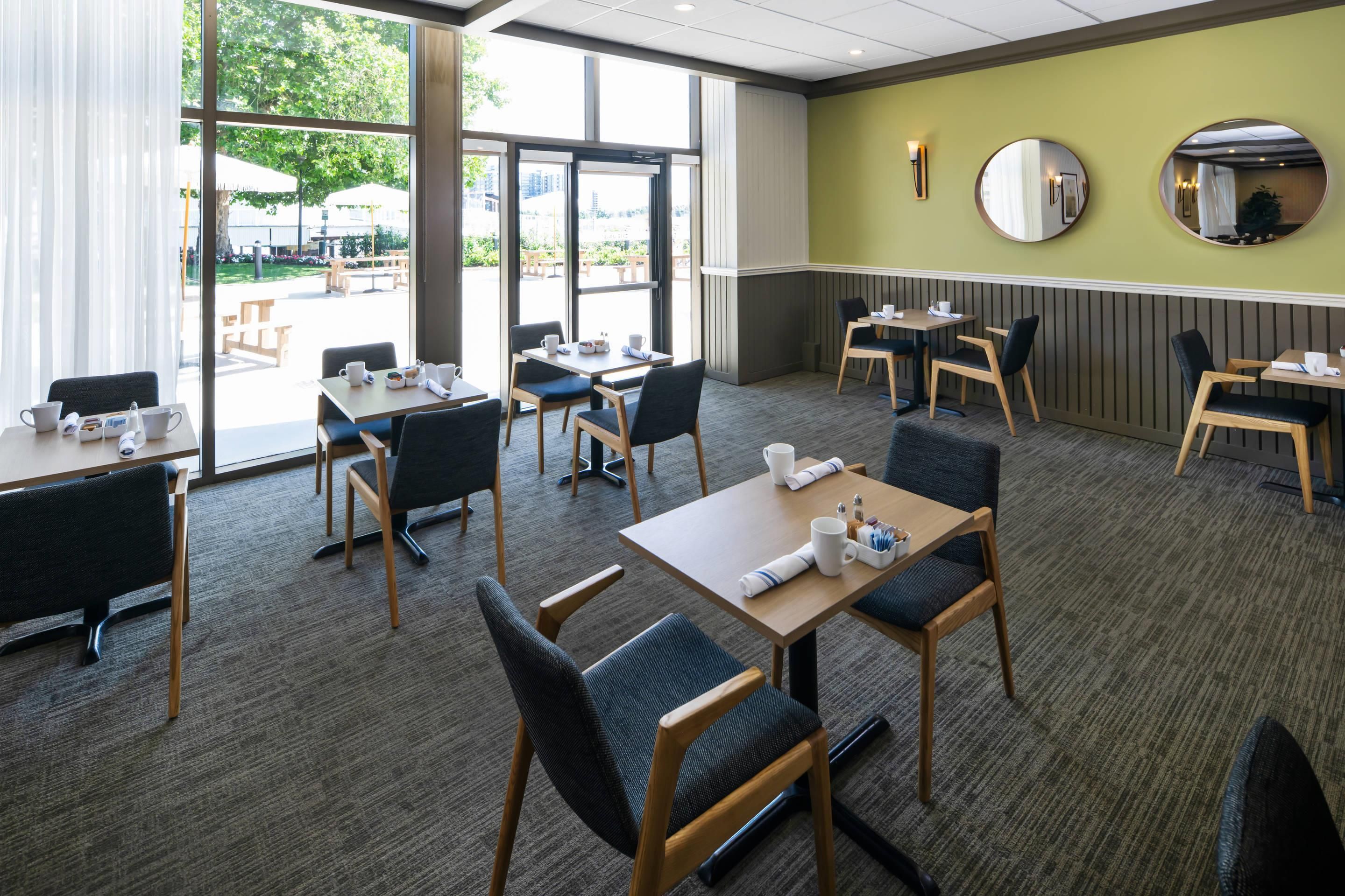 A hotel dining area with multiple tables and chairs, large windows, and a view of the outdoors.
