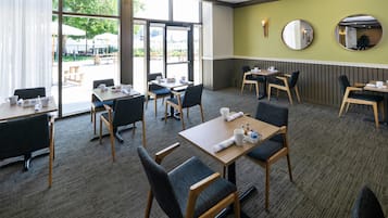 Área de jantar de hotel com várias mesas e cadeiras, grandes janelas e vista para o exterior.