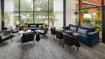 Un área de estar moderna con un conjunto de sofá azul, sillas negras y mesas redondas. Amplias ventanas ofrecen vistas al exterior.