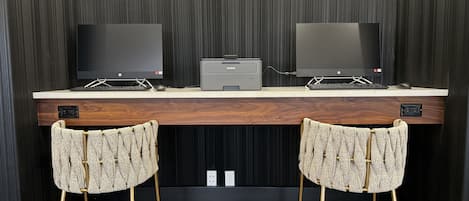 A modern workspace with two monitors, a printer, and a wooden desk. There are two upholstered chairs with gold legs.