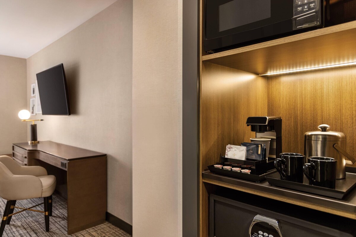 A hotel room with a desk, a chair, a flat-screen TV, a coffee station with a coffee maker, cups, and a tray.