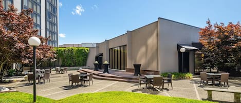 An outdoor patio area with tables and chairs, a modern building with large windows, and a clear blue sky.