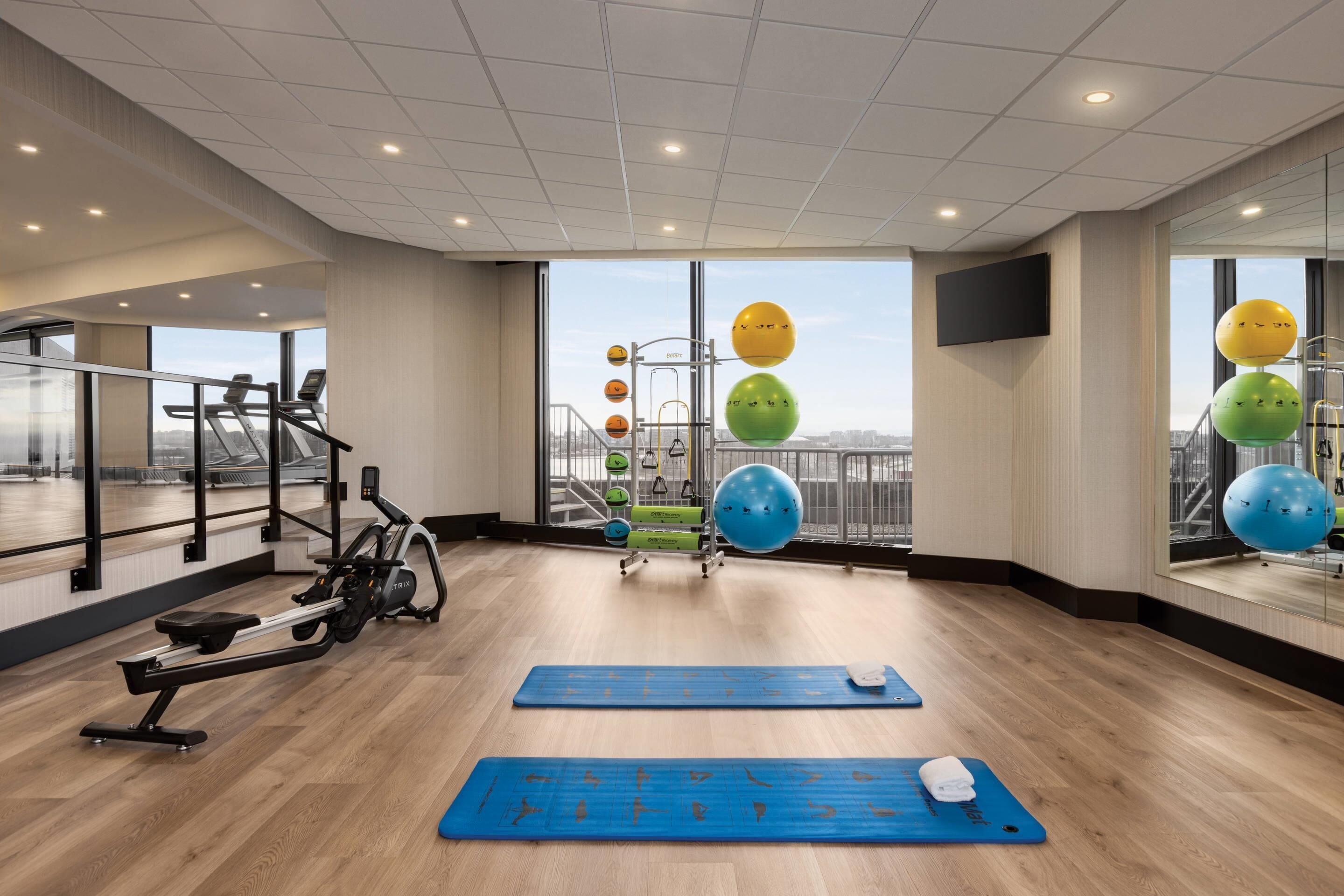 A modern gym with cardio equipment, exercise mats, and a view of the city through large windows.