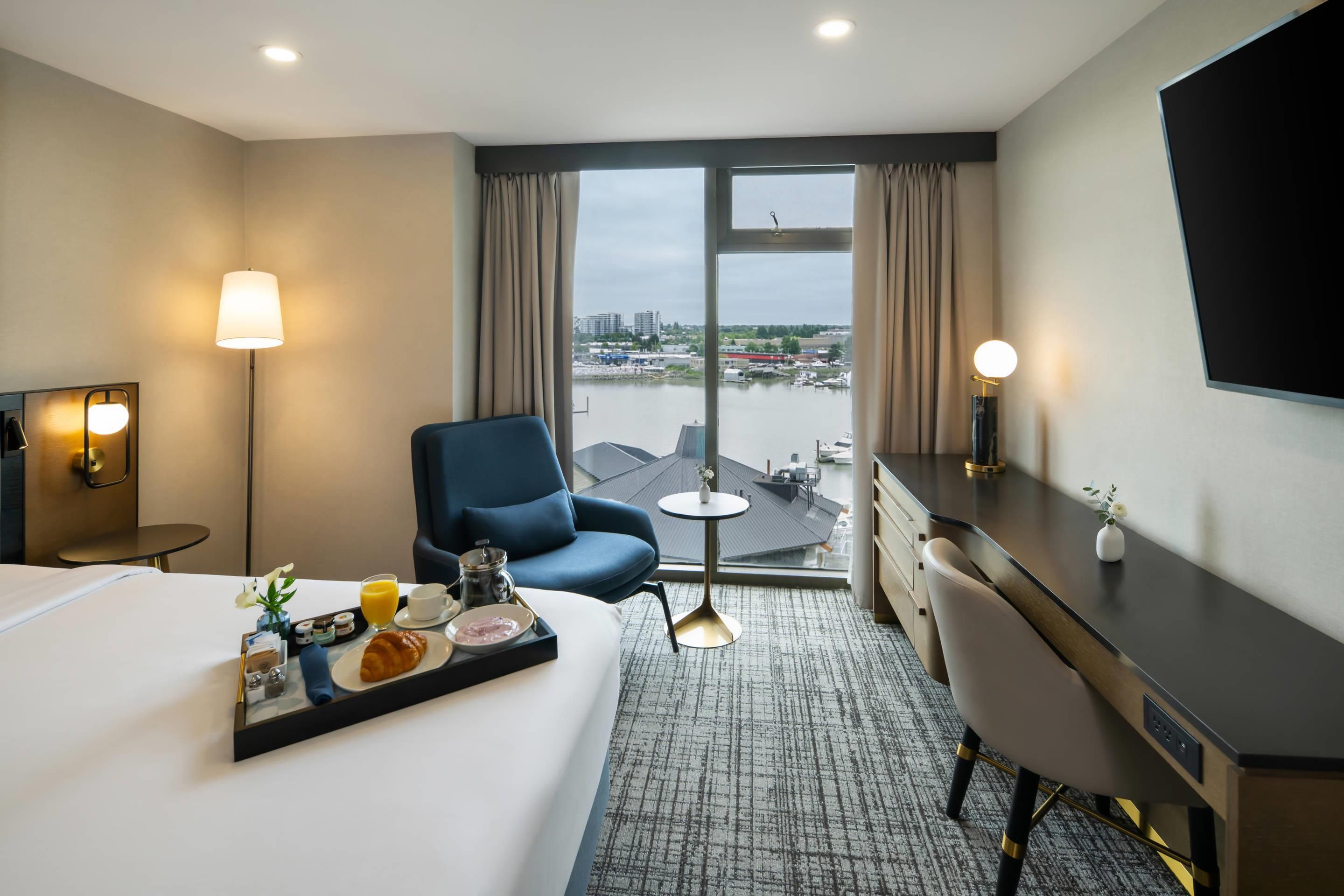 A hotel room with a bed, a desk, a chair, a small table, and a view of a body of water and buildings.