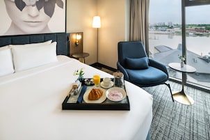 Habitación de hotel con cama, una mesa para desayunar, una silla, una lámpara y vistas a la ciudad.