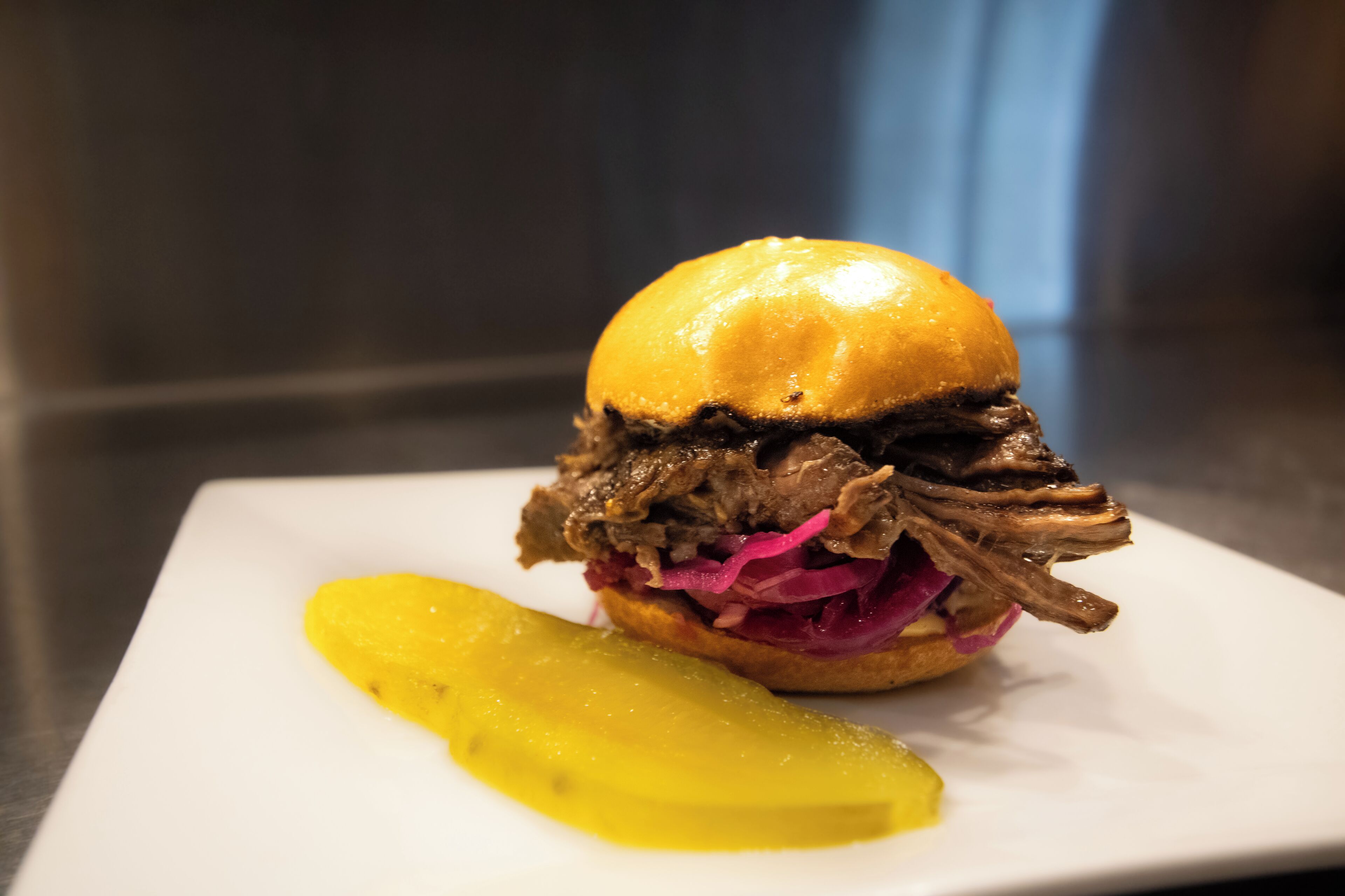 Un sandwich garni de viande effilochée et de chou rouge, servi dans un pain, accompagné d’un cornichon sur une assiette blanche.