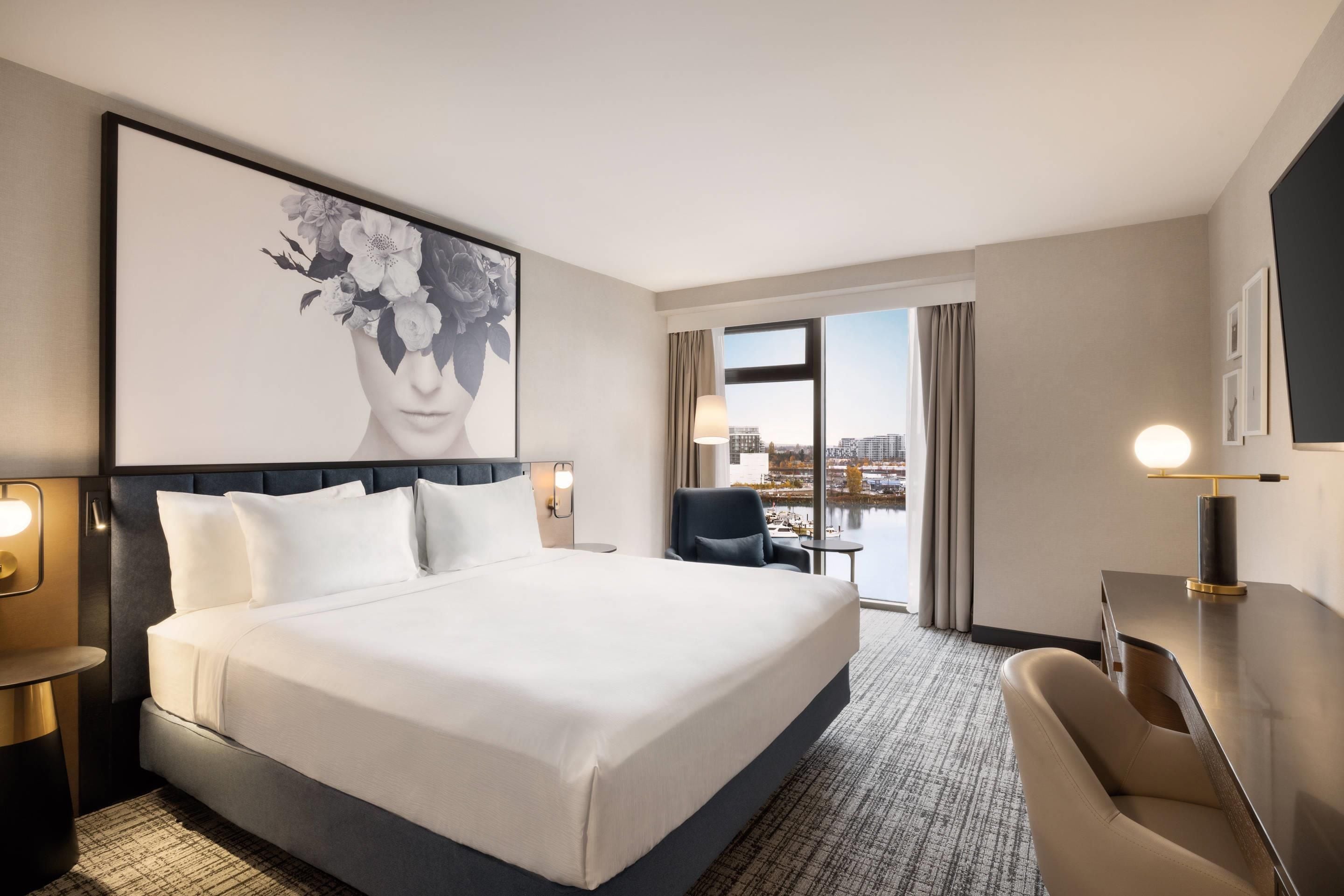A hotel room with a large bed, a desk, a chair, and a view of the city through the window.