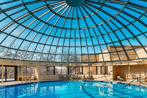 A swimming pool with a glass dome ceiling, lounge chairs, and a view of trees outside.