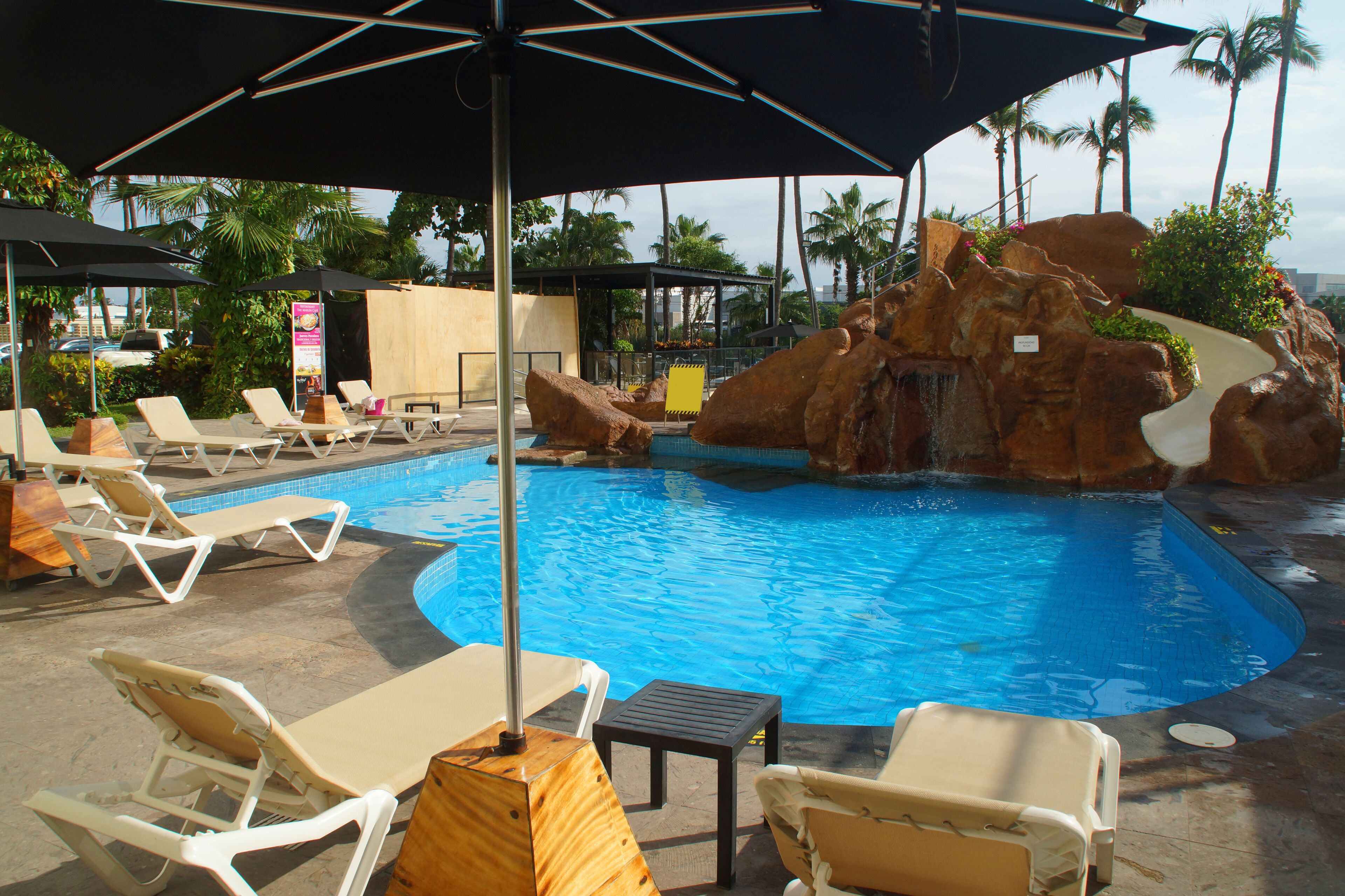 A pool area with lounge chairs and umbrellas, surrounded by palm trees and a rocky landscape.