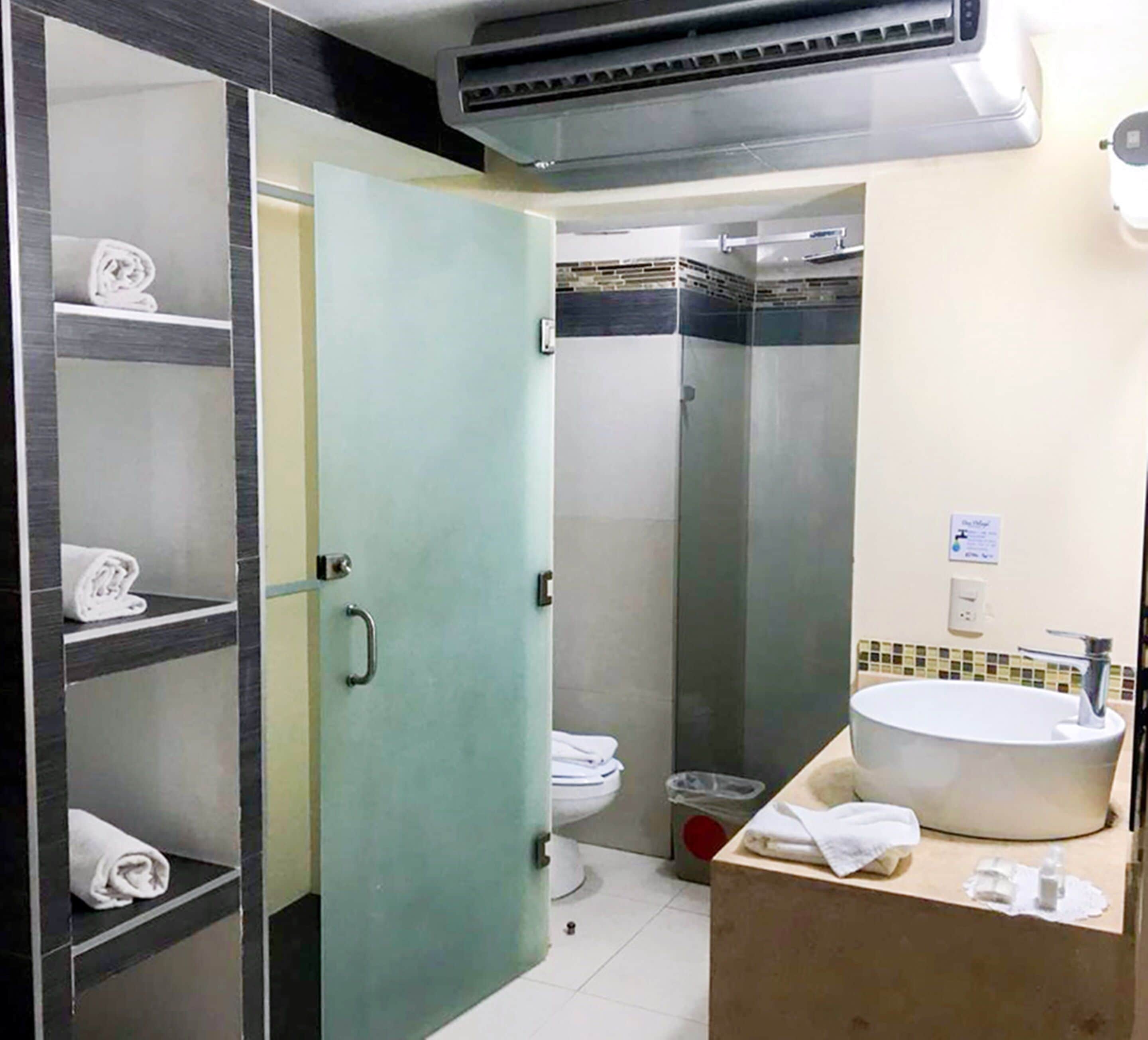 Une salle de bain avec des toilettes, un lavabo et une douche. Il y a des serviettes sur les étagères et une poubelle.