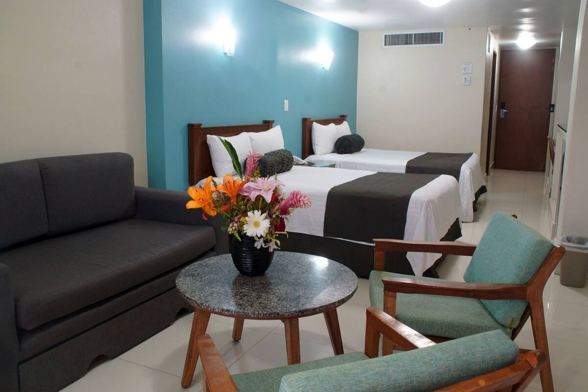 A hotel room with a sofa, two single beds, a small table with a flower vase, and a chair.