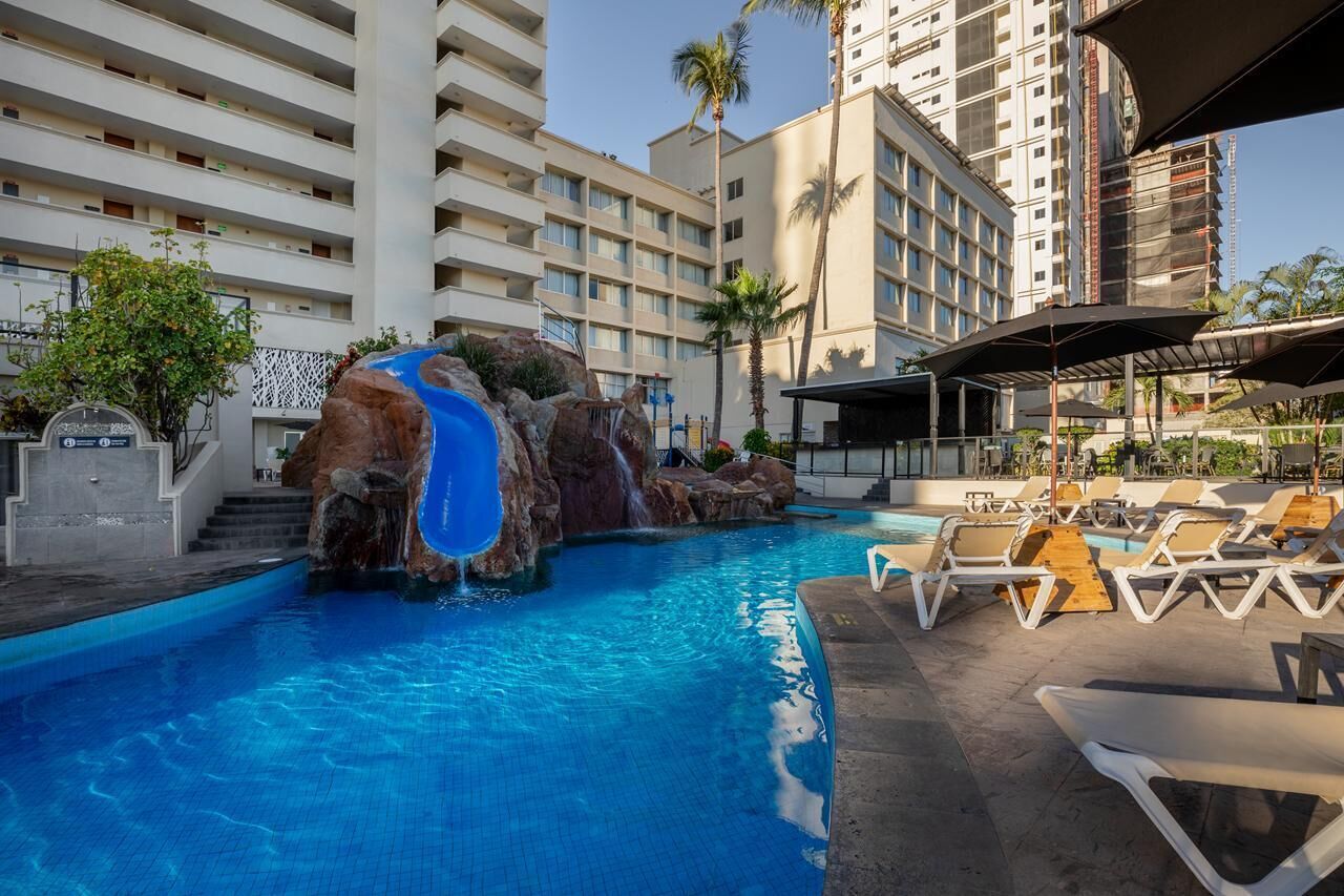 A hotel pool with a waterfall feature, lounge chairs, and a multi-story building in the background.