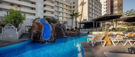 A hotel pool with a waterfall feature, lounge chairs, and a multi-story building in the background.