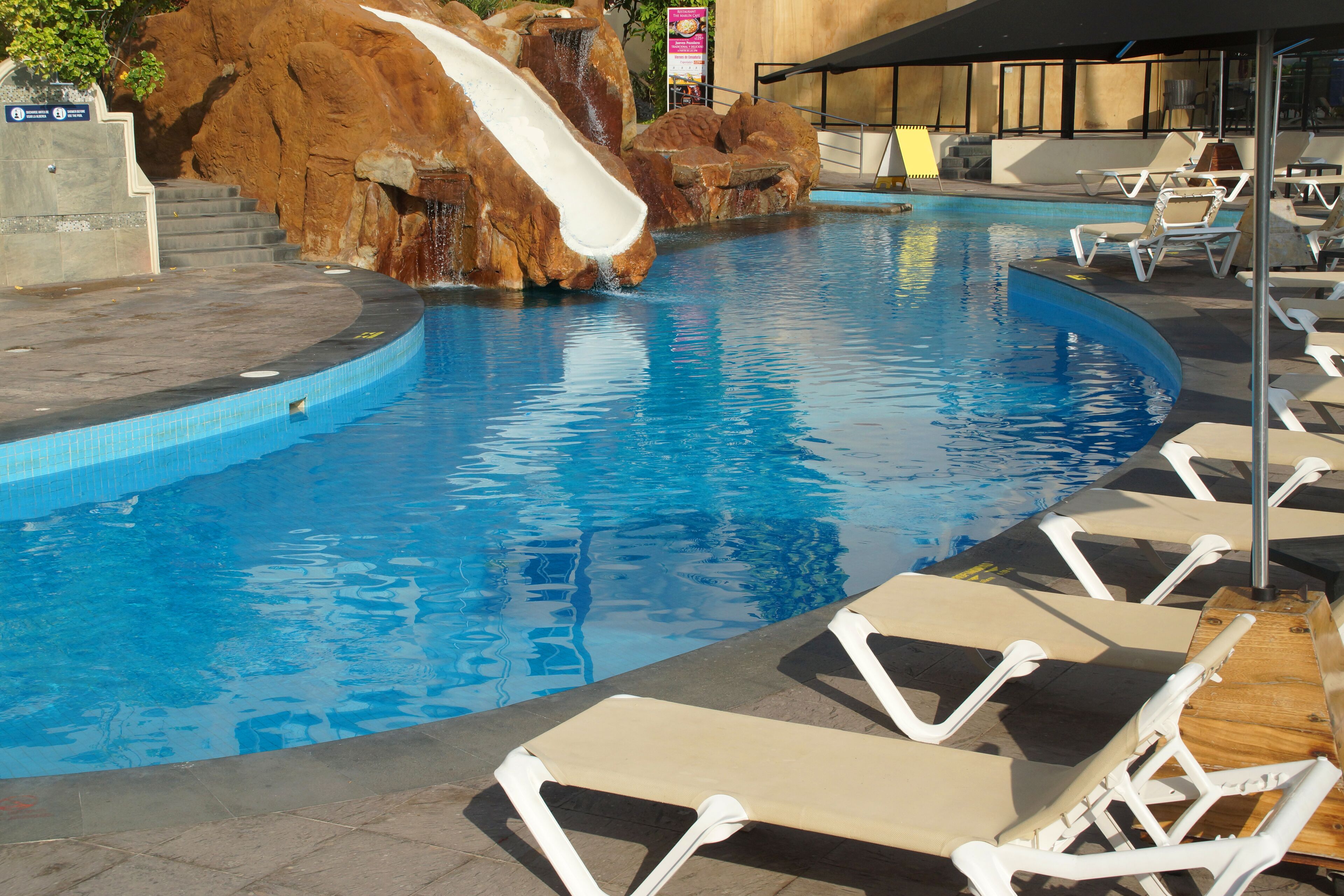 A swimming pool with a water slide, lounge chairs, and a rock formation.