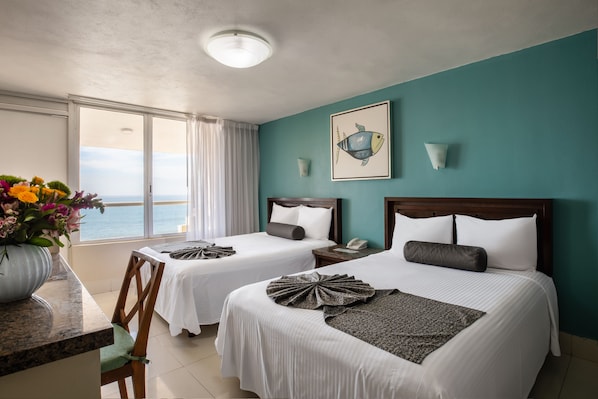 A hotel room with two beds, a large window with a view of the sea, a vase of flowers, and a framed fish artwork on the wall.