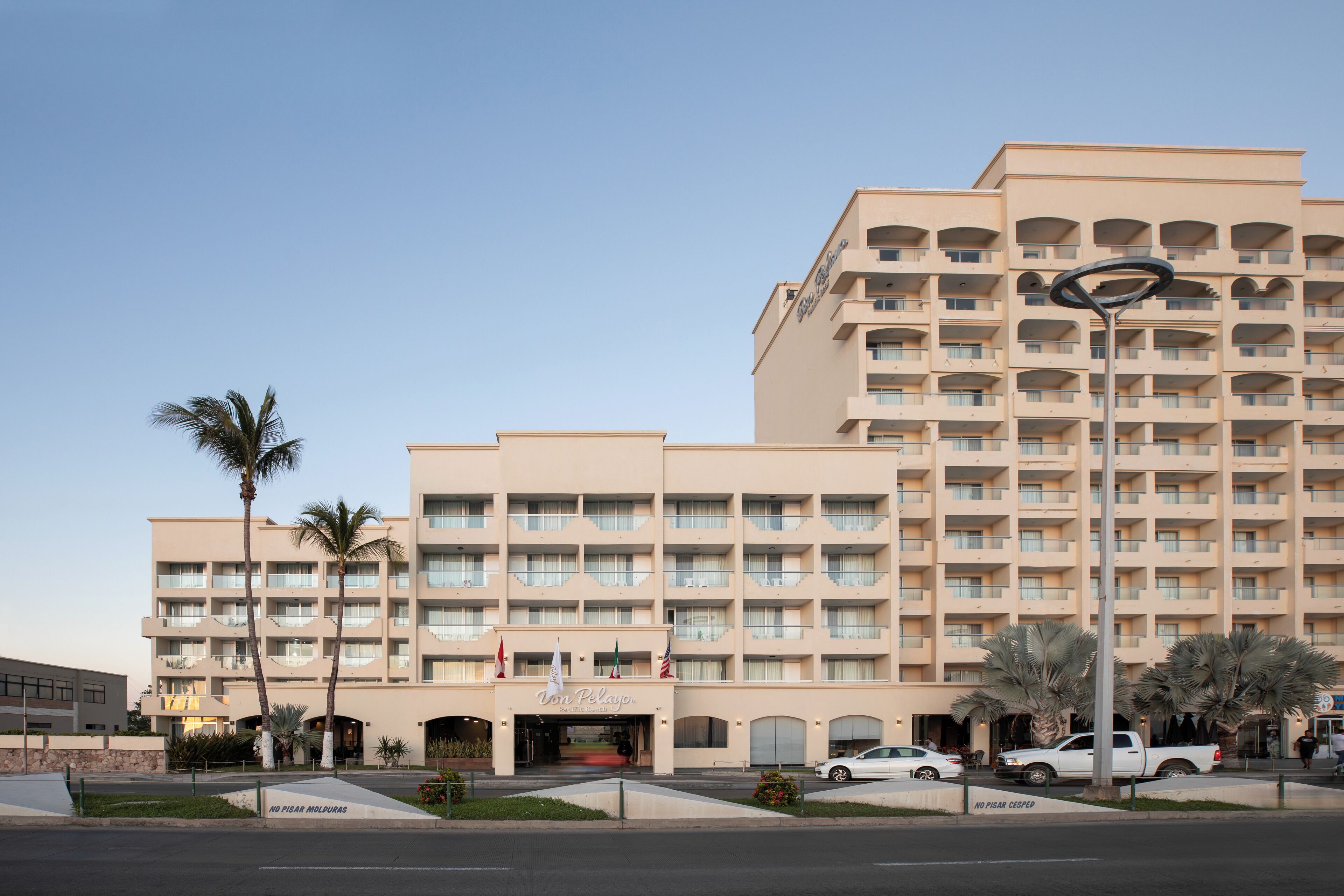 Um hotel de vários andares com entrada central, palmeiras e estacionamento.