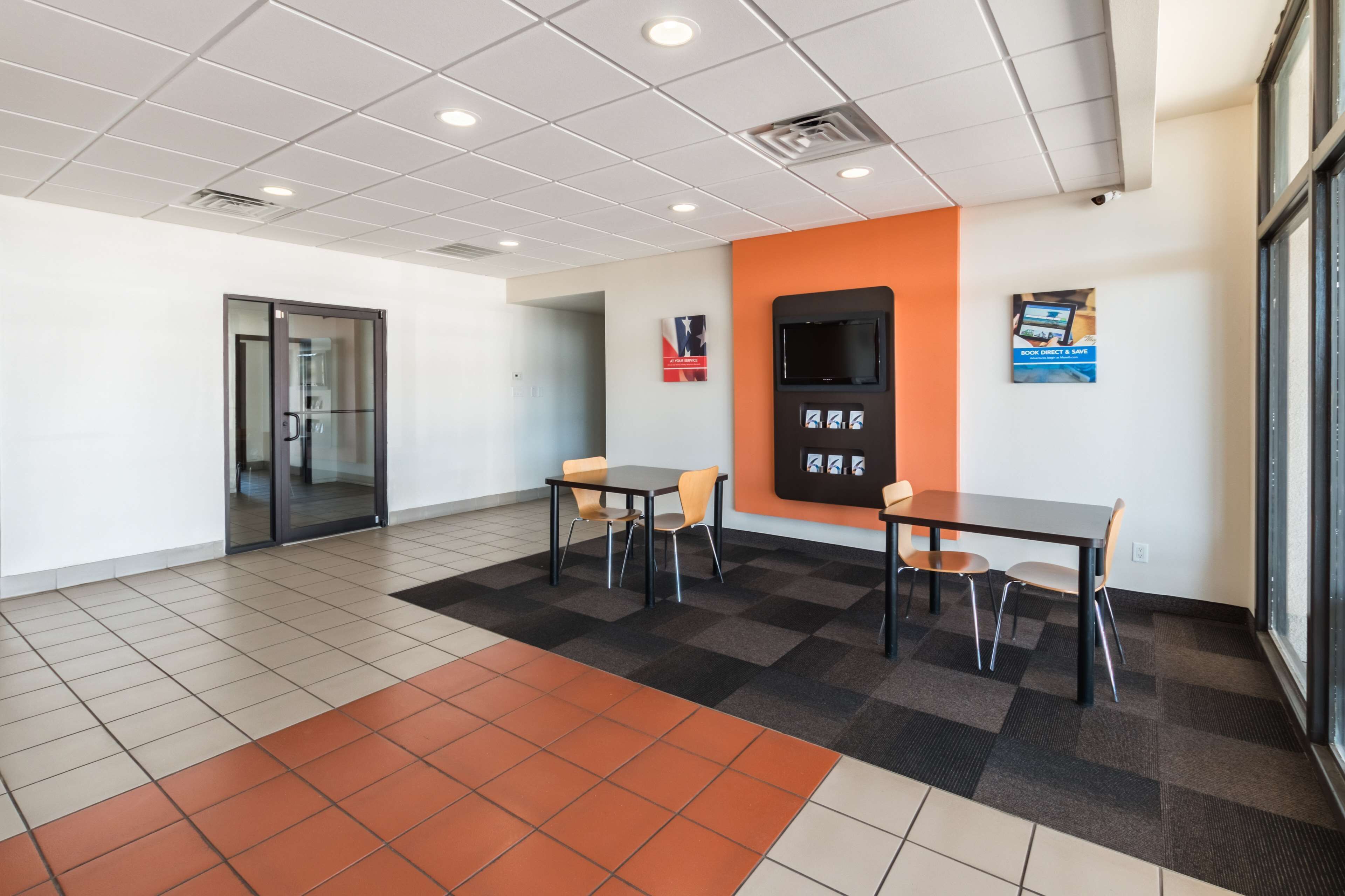 A modern office space with a meeting room, two tables with chairs, a large window, and a wall-mounted screen.