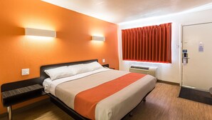 A hotel room with a bed, bedside table, wall-mounted light, window with curtains, and a door. - Motel 6 Stephenville, TX (Stephenville)