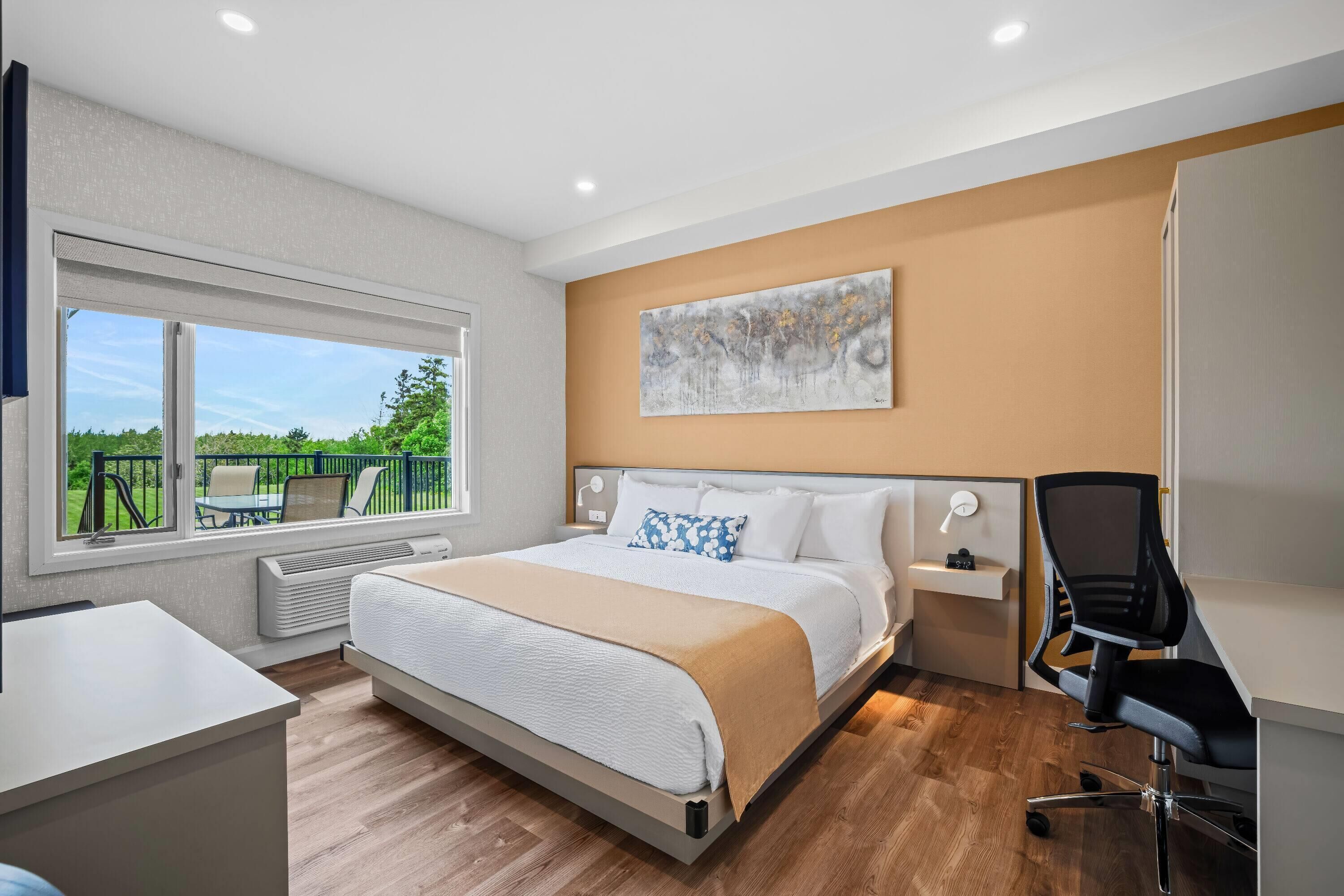 A modern hotel room with a large bed, a desk, and a chair. There is a window with a view of greenery outside.