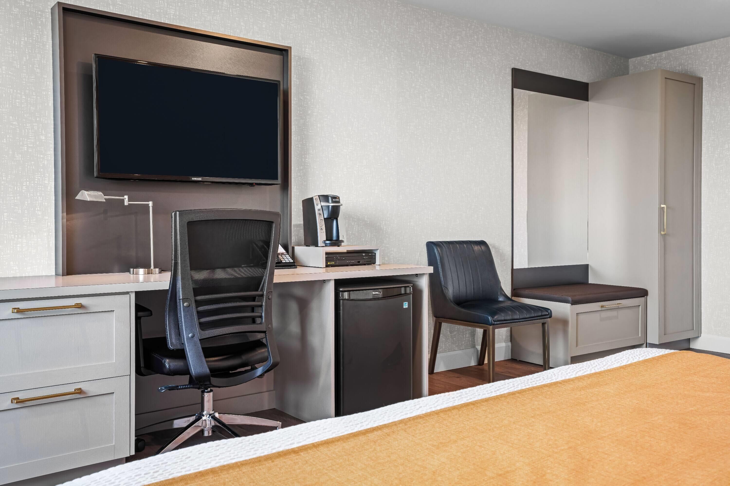 A hotel room with a bed, a desk with a chair, a coffee machine, a TV, and a wardrobe.