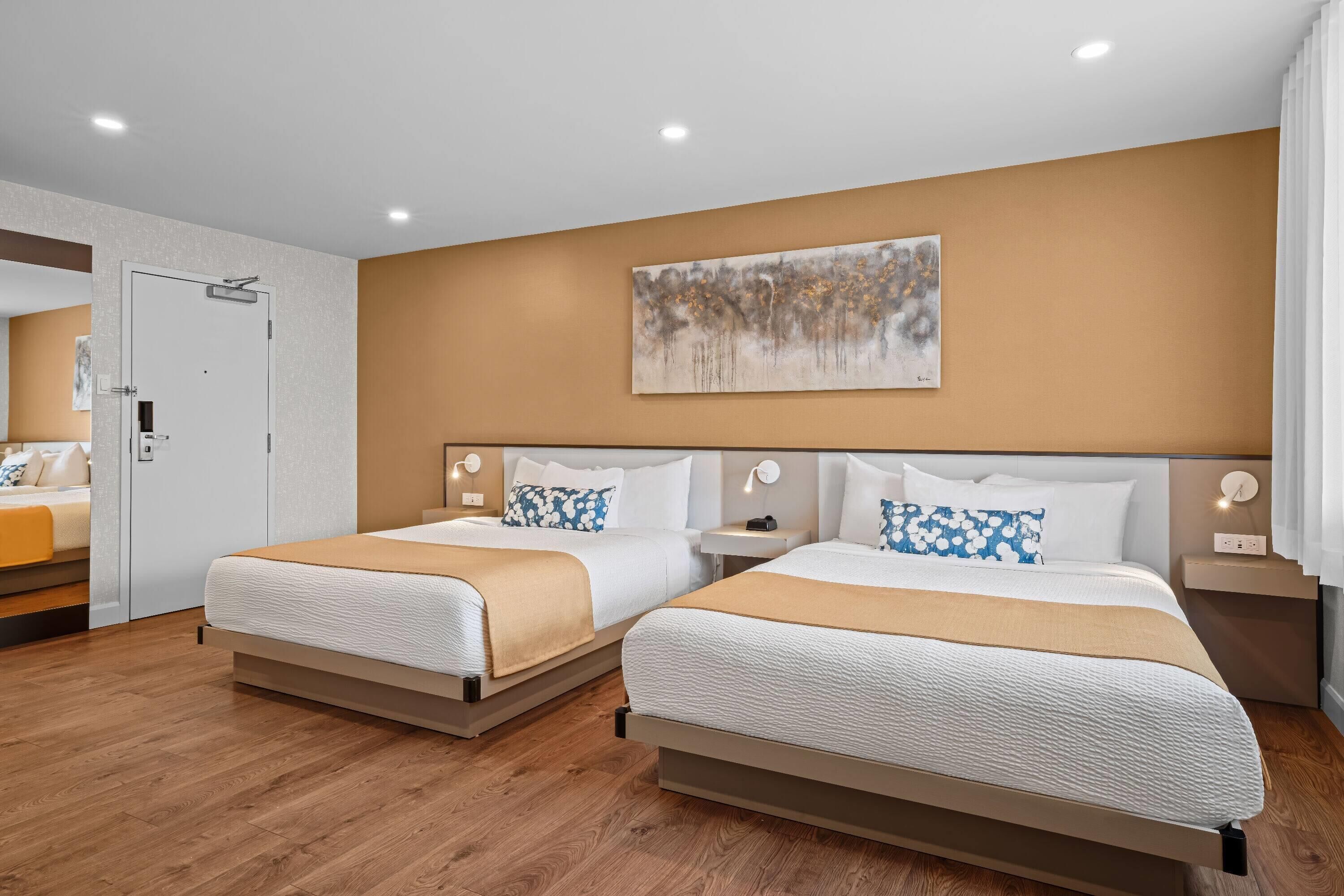 A hotel room with two beds, wooden flooring, a painting on the wall, and a door to the left.