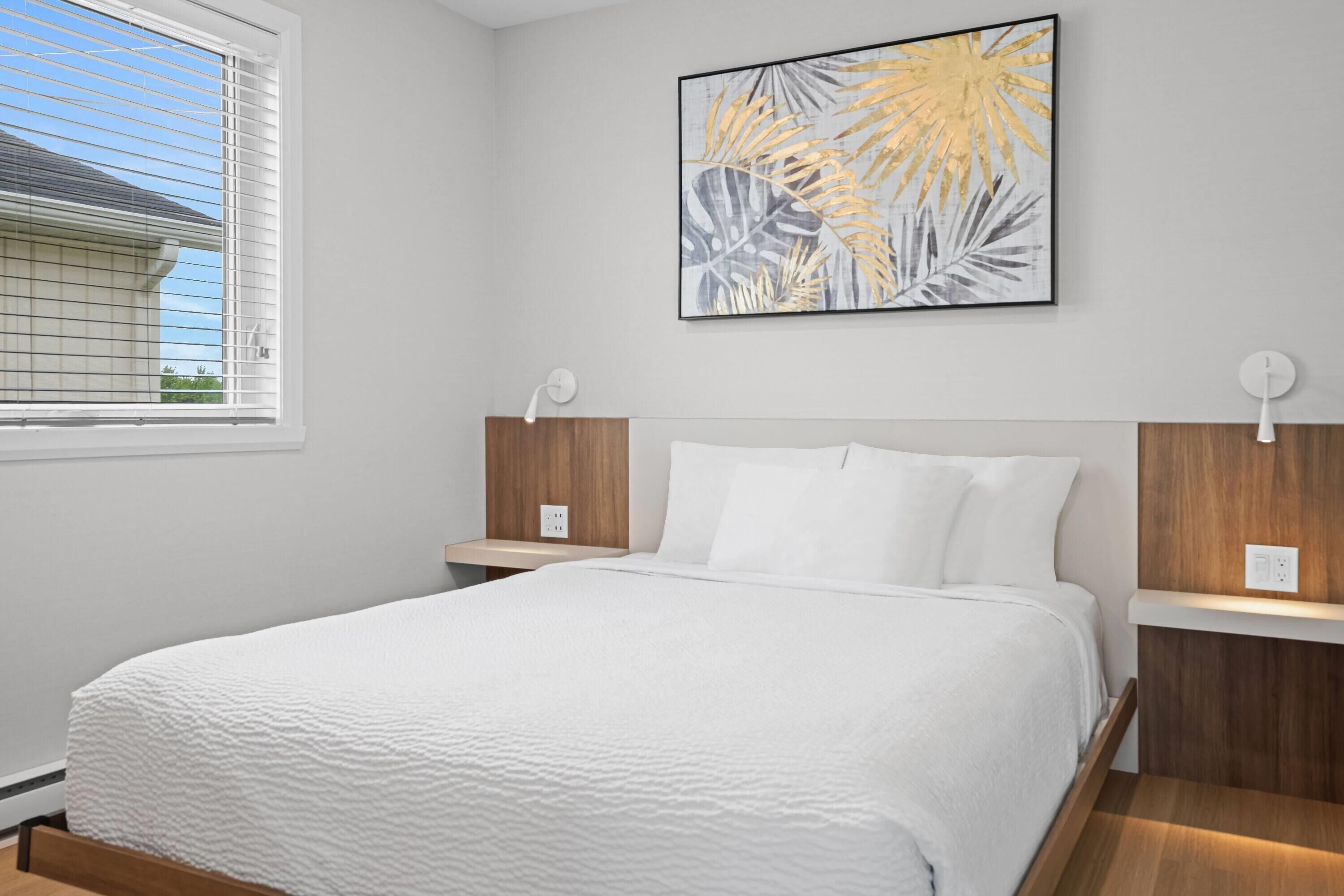 A bedroom with a bed, white bedding, a wall-mounted artwork, bedside lamps, and a window with blinds.