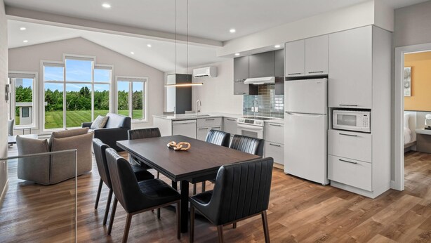 A modern kitchen with white cabinetry, a dining area with a wooden table and black chairs, and large windows offering a view of greenery. - Days Inn by Wyndham Riviere-Du-Loup (Rivière-du-Loup)