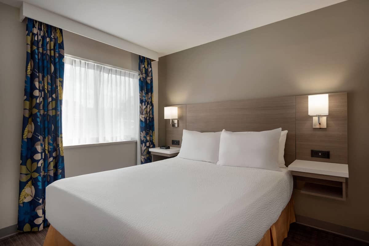 A hotel room with a large bed, two bedside tables, a nightstand, and a window with sheer curtains.