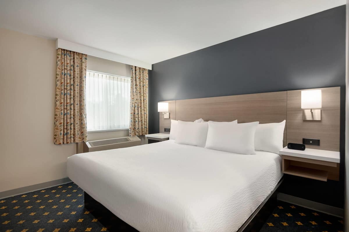 A hotel room with a large bed, two bedside tables with lamps, a window with curtains, and a dark wall.