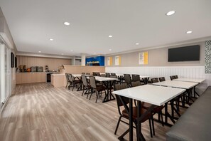 Un espace repas moderne avec des tables et des chaises blanches, une cuisine en arrière-plan et un panneau indiquant : « Le petit-déjeuner est offert ».