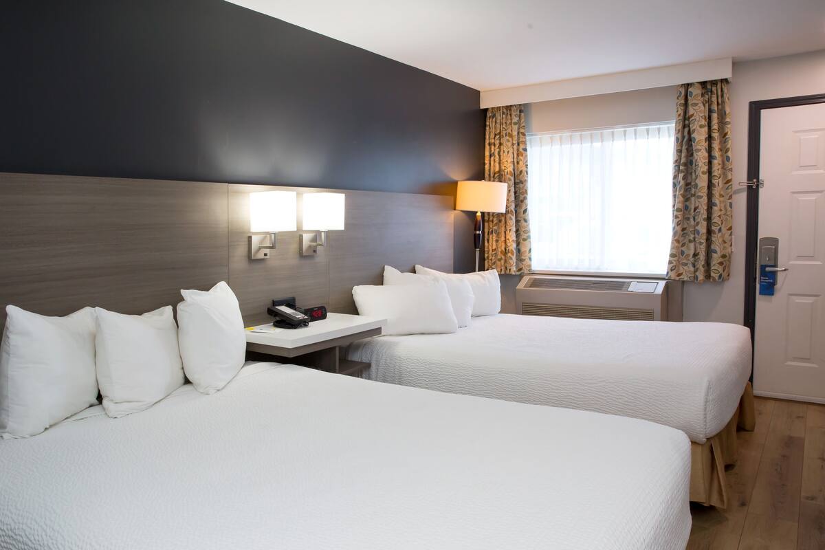 A hotel room with two beds, a bedside table with a lamp, a window with curtains, and a door.