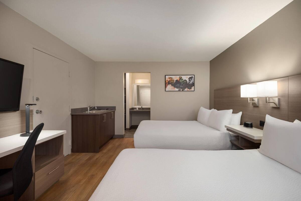 A hotel room with two beds, a desk, a chair, a TV, a sink, and a mirror.