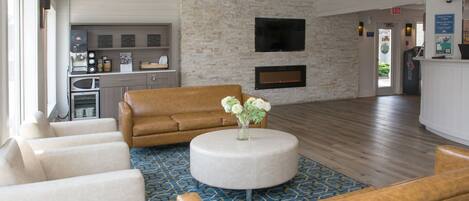 A spacious living area with a brown sofa, white armchairs, and a round coffee table with flowers. A kitchen with appliances and a flat-screen TV is visible in the background.