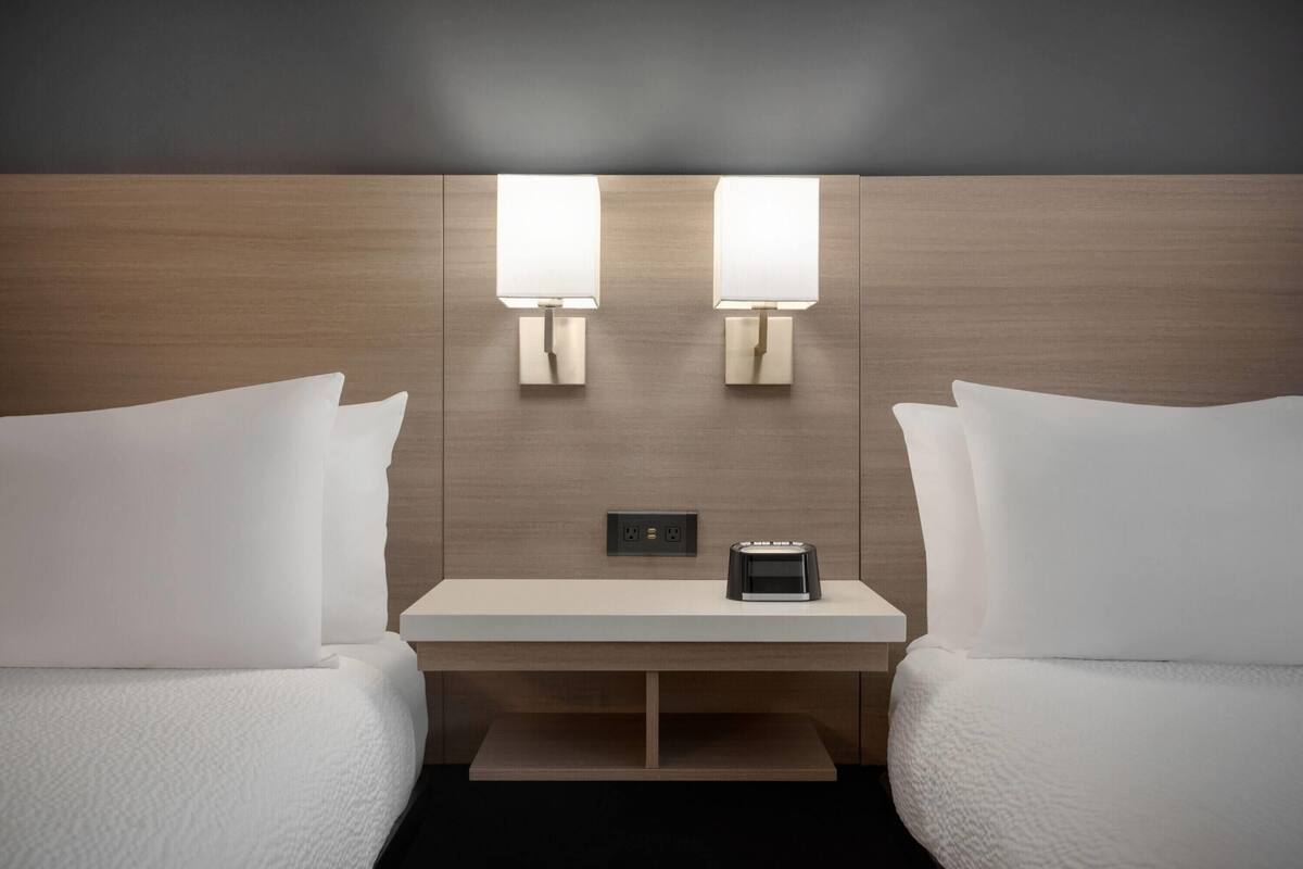 A hotel room with two beds, a bedside table, two wall-mounted lamps, and a small table with a clock.
