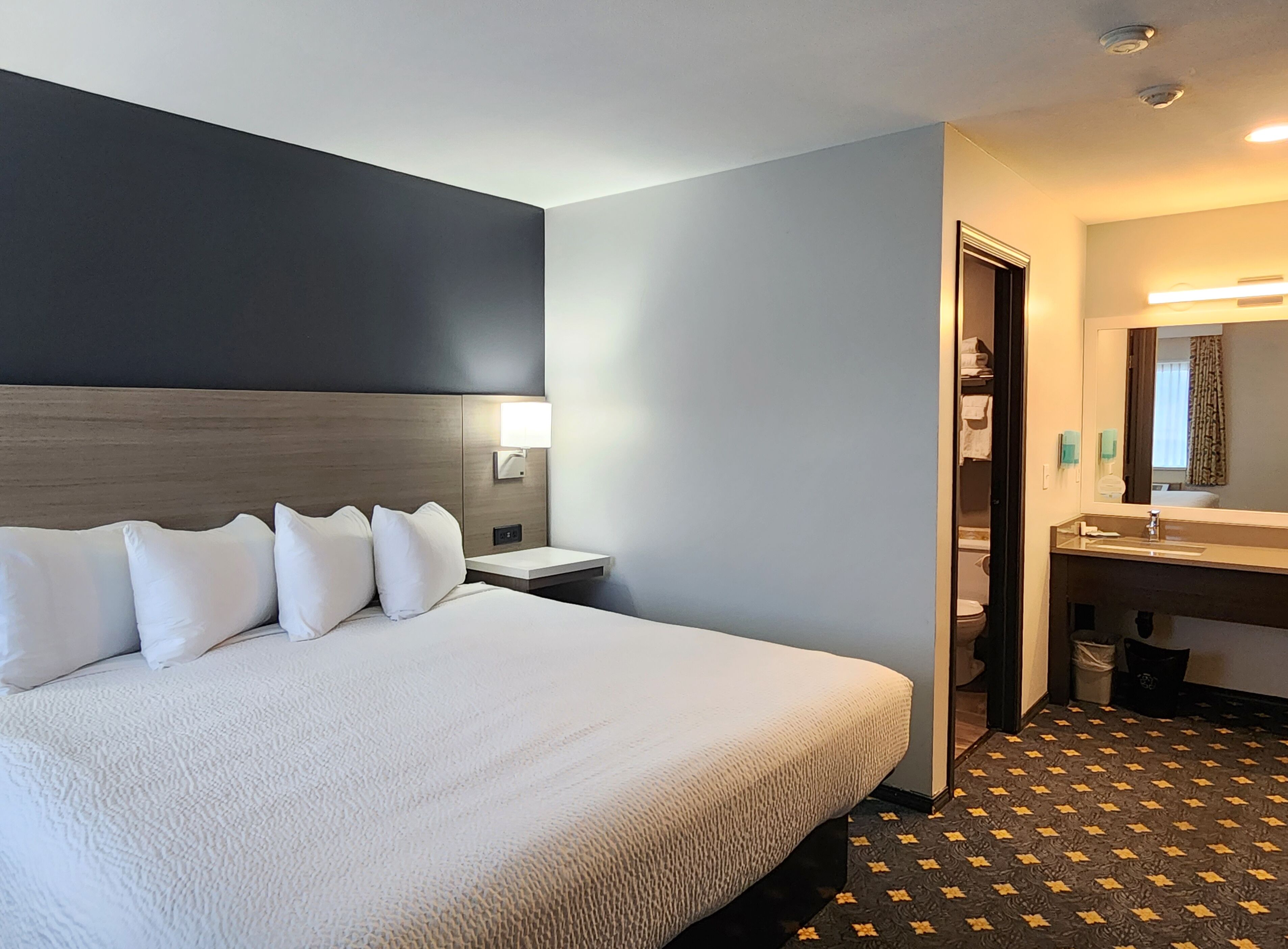 A hotel room with a large bed, a nightstand, a wall-mounted lamp, a bathroom with a sink and mirror, and a patterned floor.