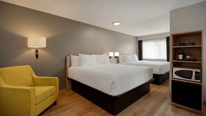 Habitación de hotel con cama, una silla, una mesita de noche, lámpara, estante con microondas y ventana con cortinas.