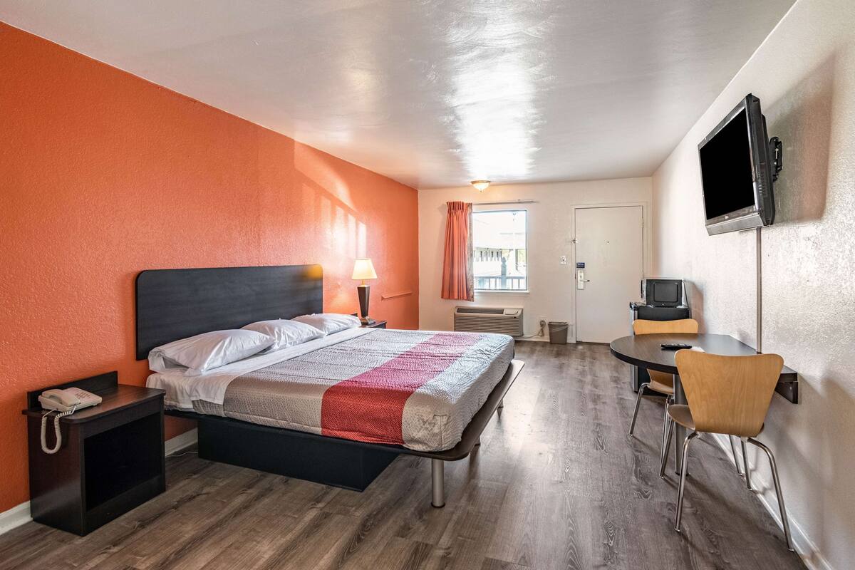A hotel room with a bed, bedside table with a phone, a desk with a chair, a TV mounted on the wall, and a window with curtains.