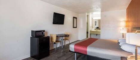 Habitación de hotel compacta con cama, escritorio con microondas, televisor y una pequeña mesa con sillas.