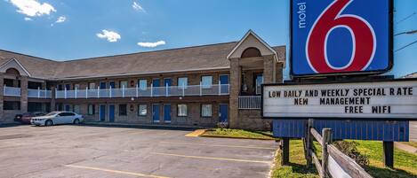A motel with a large sign displaying the number 6. It has a parking lot with several cars, a brick building with multiple windows, and a signboard advertising low rates and free WiFi.