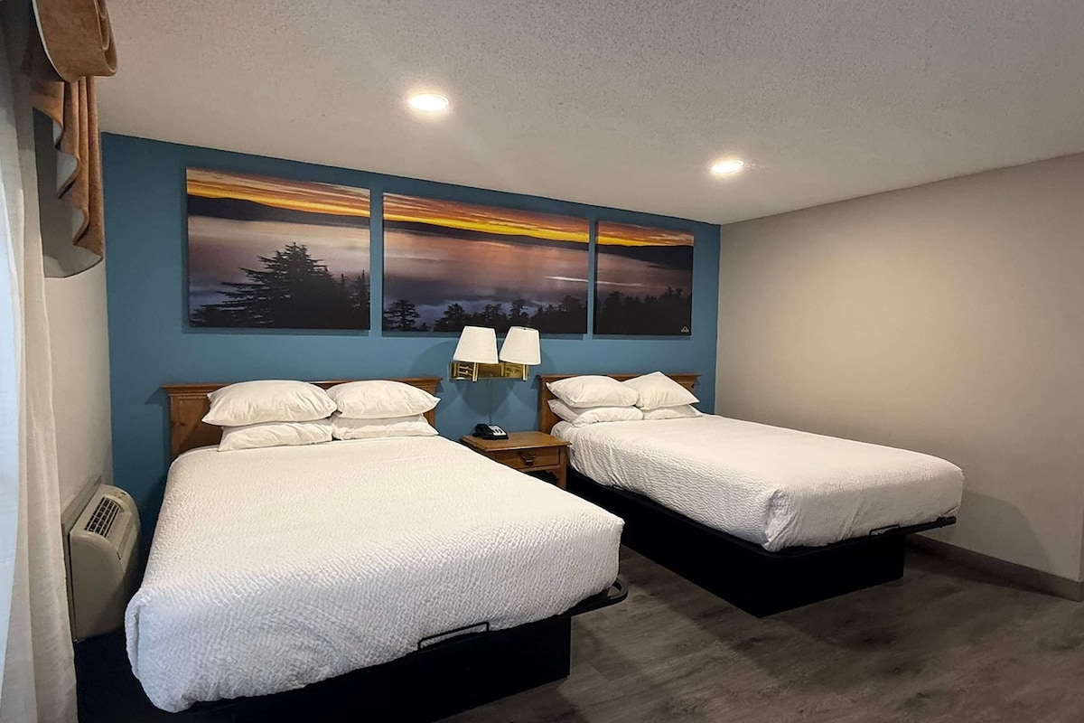 A room with two beds, a nightstand with a lamp, a wall-mounted painting, and a ceiling light.