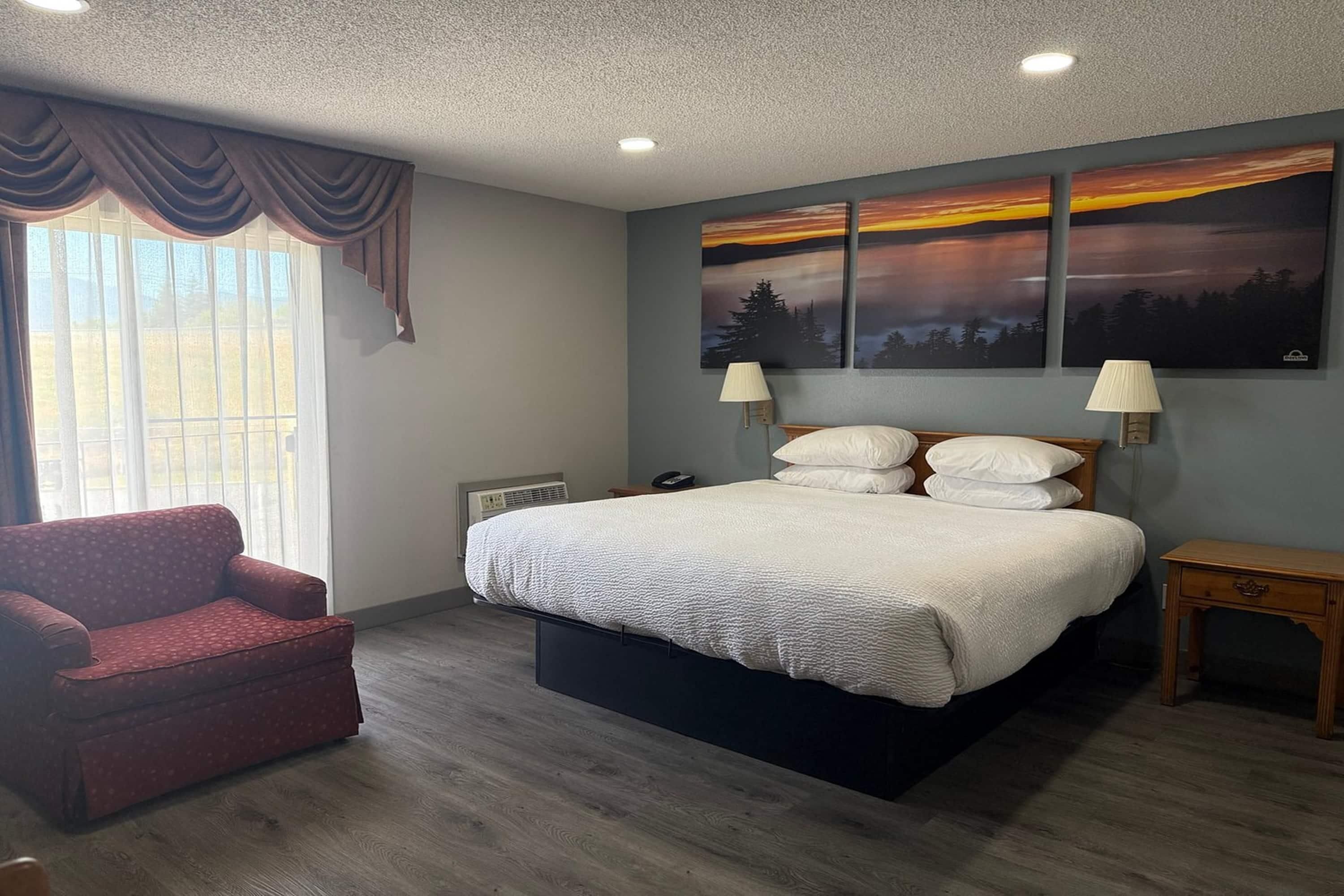A bedroom with a bed, a chair, a bedside table, a lamp, and a painting on the wall.